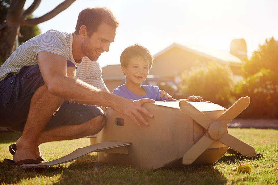 About Our Agency - Smiling Father and Son Playing Outside in the Backyard with a Cardboard Plane on a Warm Summer Afternoon